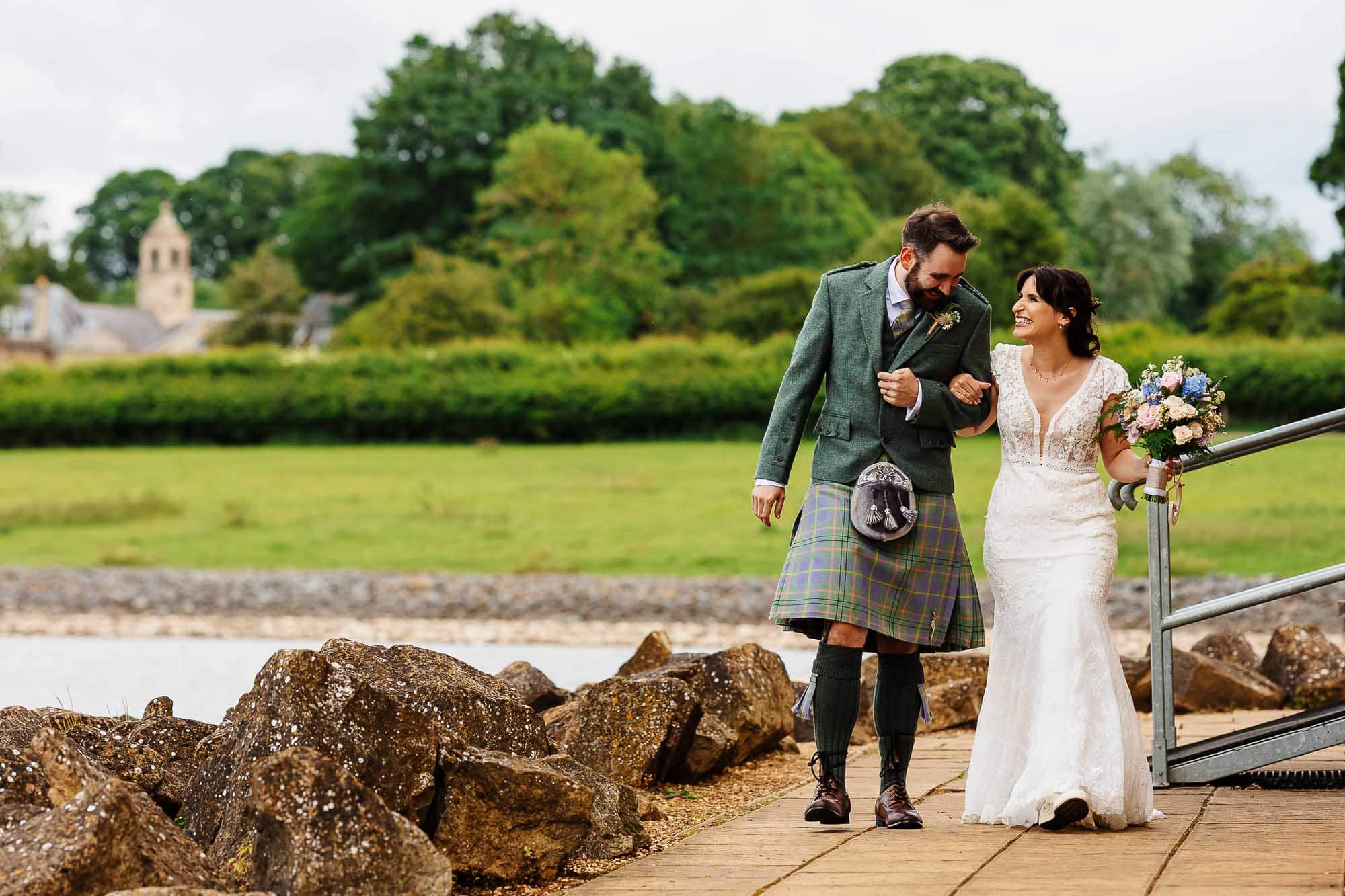 A bride and groom walking along a lakeside path in traditional Scottish attire, surrounded by lush greenery and scenic landscape.