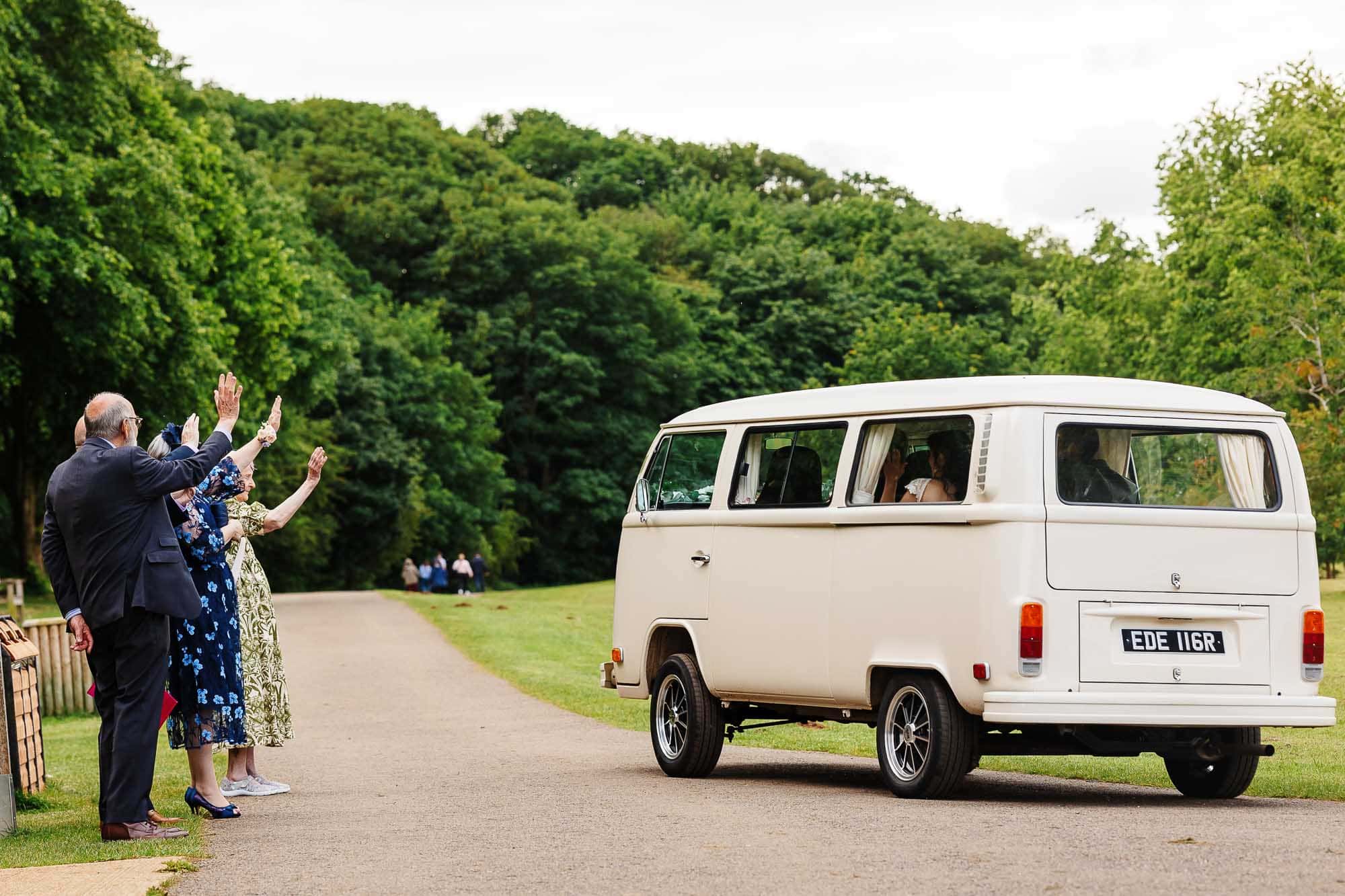 Classic white wedding van with happy guests waving farewell on a scenic outdoor wedding day.