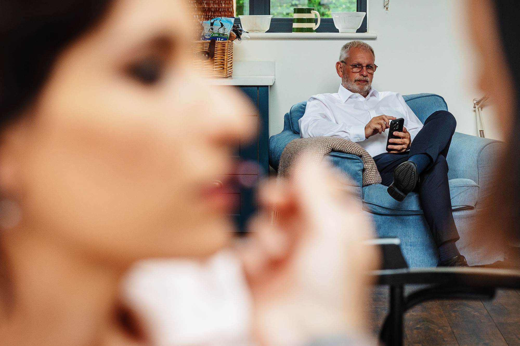 Wedding preparation at BGS Weddings, groom sitting on a blue sofa, checking his phone, with a blurred bride in the foreground.