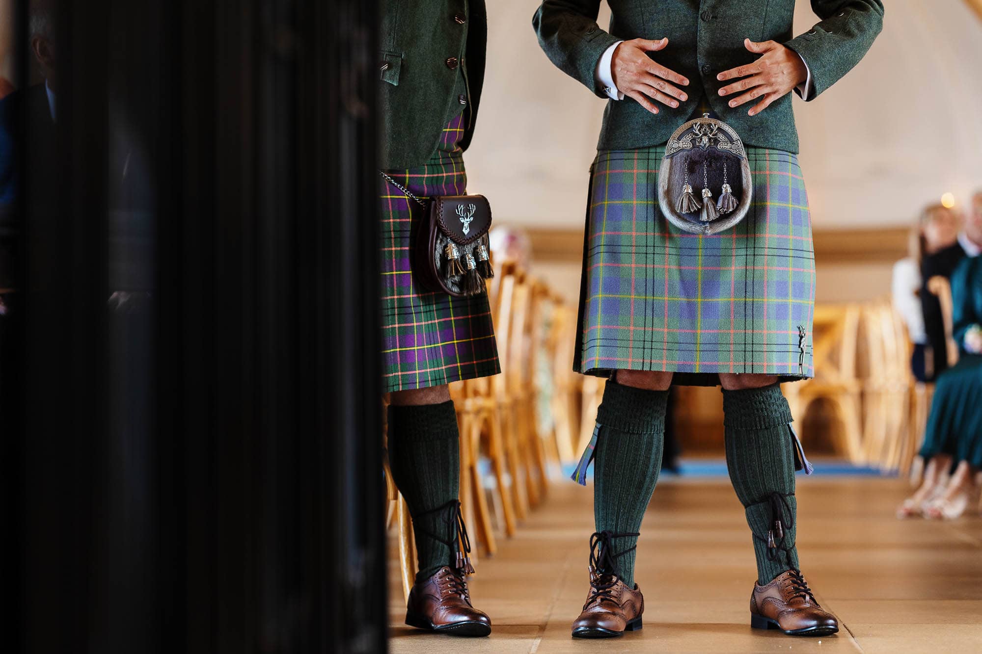 Two men in traditional Scottish kilts and jackets standing indoors during a wedding ceremony, with guests seated in the background.
