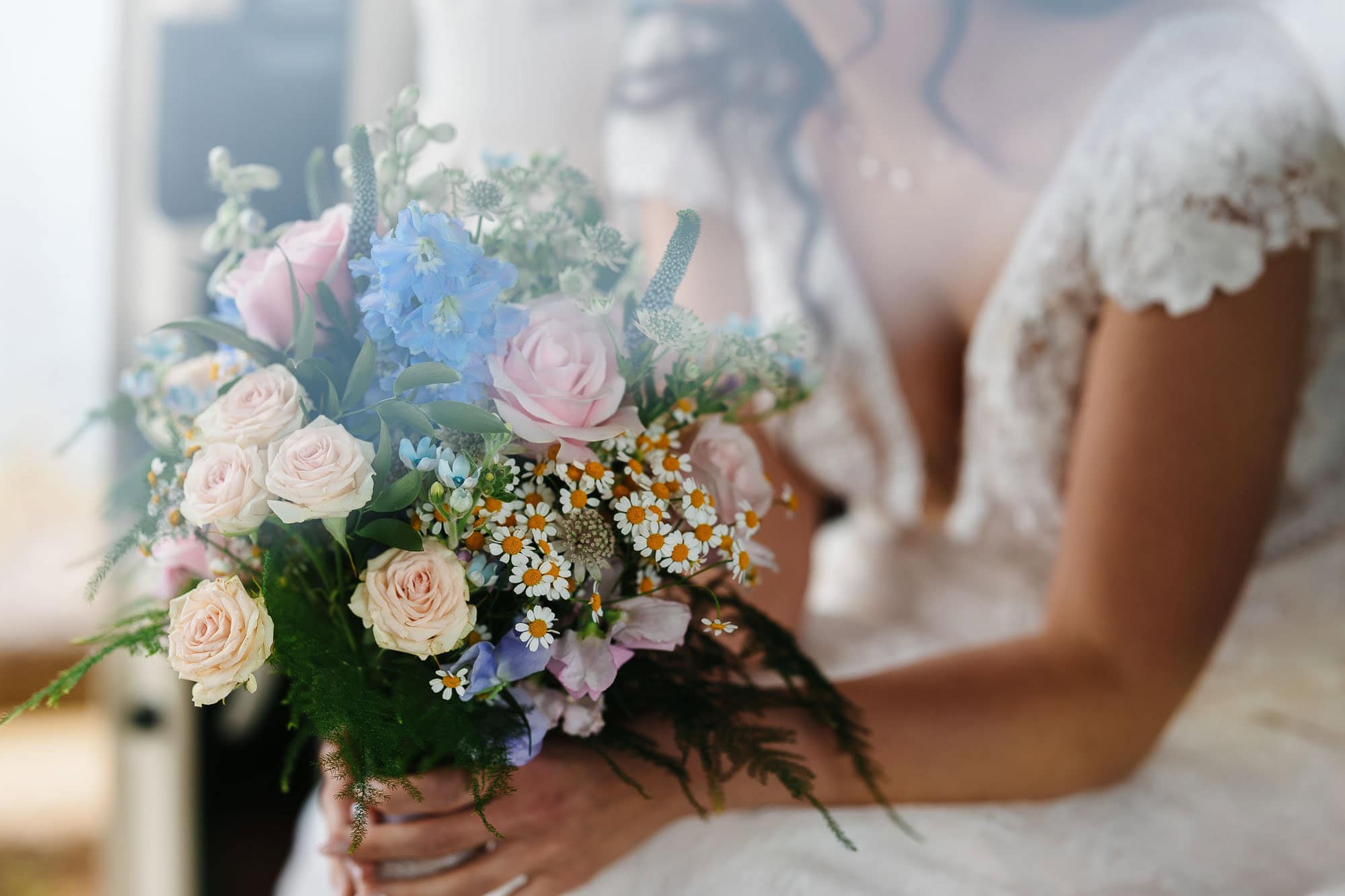 Bridal bouquet featuring pink roses, blue hydrangeas, and white daisies, held by a bride in a lace wedding dress. Perfect for wedding photography and floral arrangements.