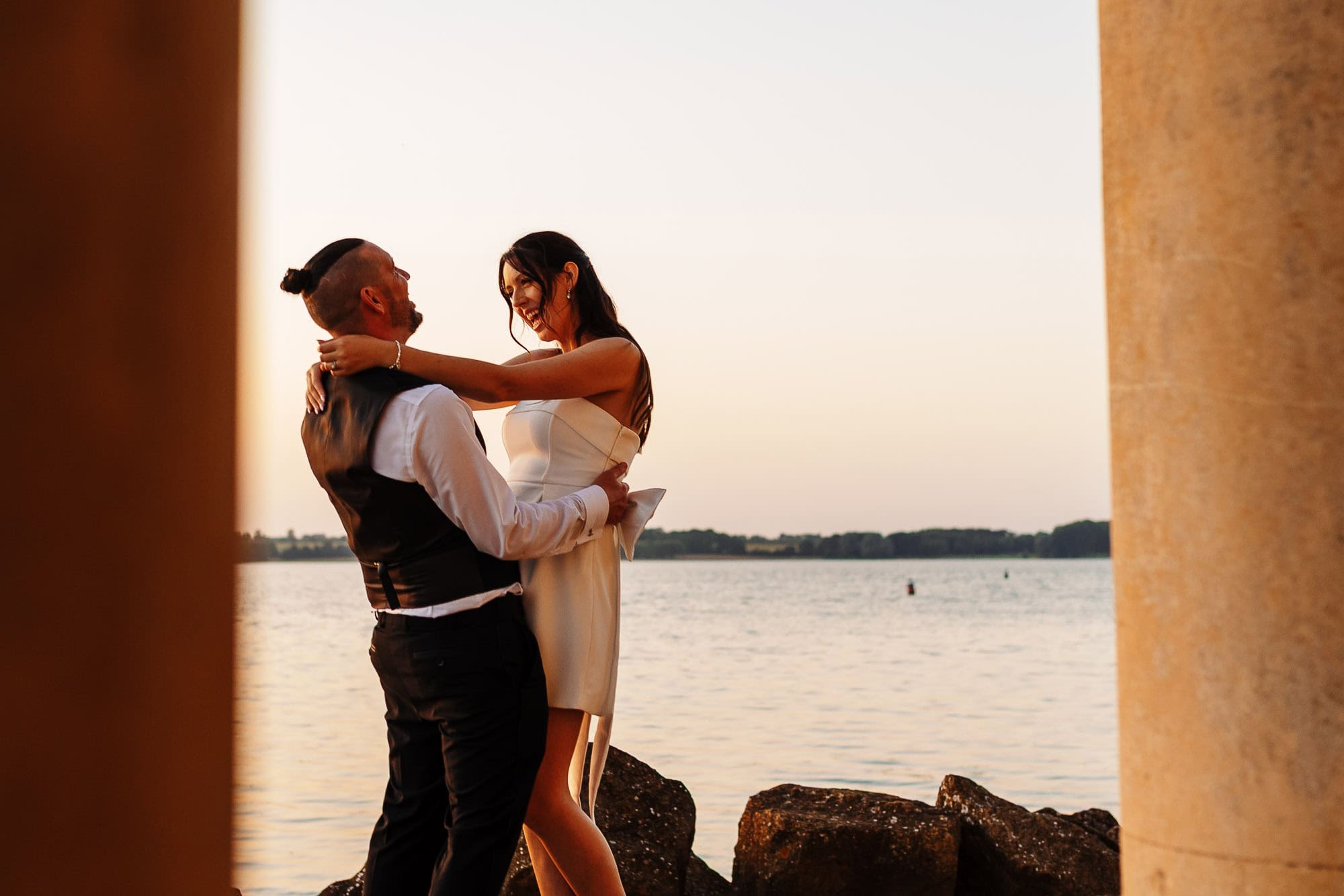 Beautiful wedding couple embracing by the water at sunset, capturing a romantic moment during their special day, with the BGS Weddings logo visible.