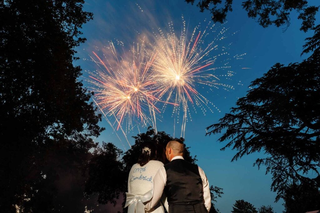 Couple watching fireworks display at night, celebrating a wedding, with trees framing the scene, capturing a romantic moment during a wedding celebration.