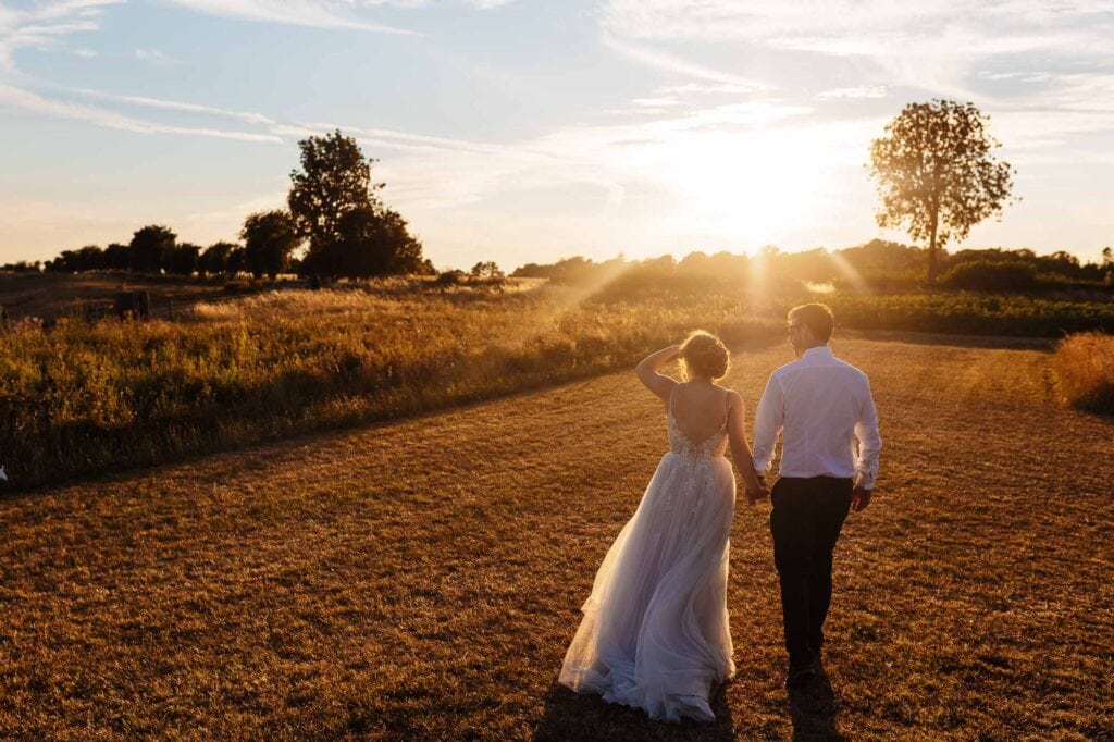 Bride and groom walking hand in hand during sunset on a wedding day, with warm golden light and scenic countryside background, capturing love and happiness.