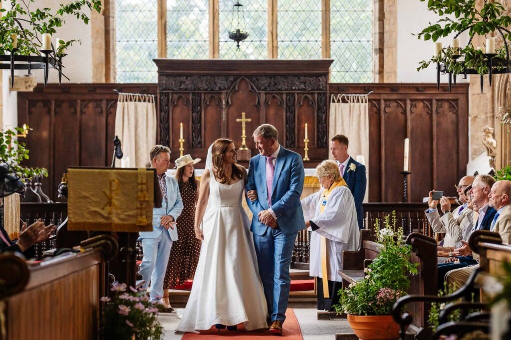Wedding ceremony at BGS Weddings in a church with bride and groom, officiant, and guests, featuring traditional decor and natural light from large windows.