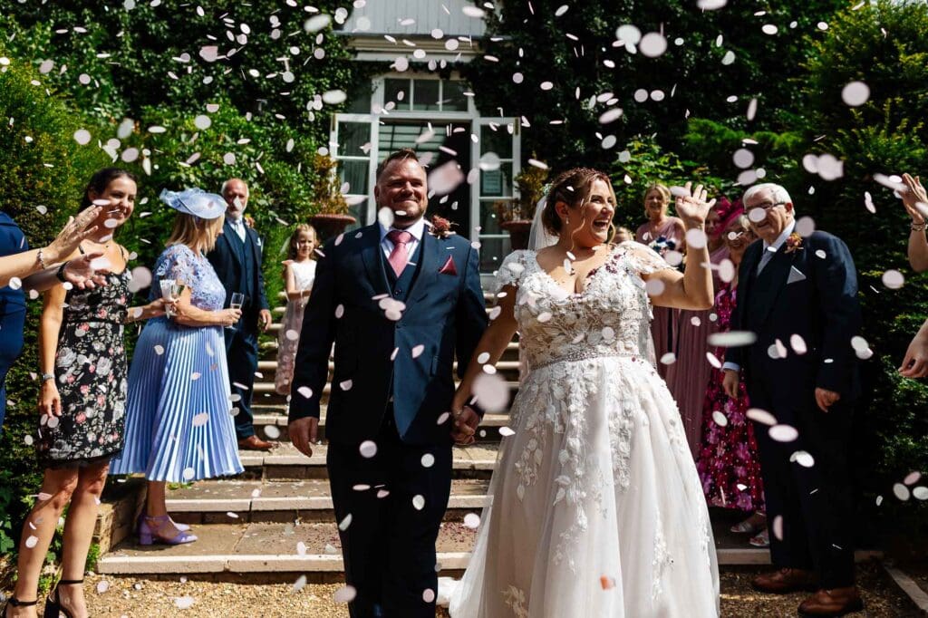 Happy newlyweds celebrating with guests outside a charming venue, surrounded by confetti and greenery, capturing a joyful wedding moment.