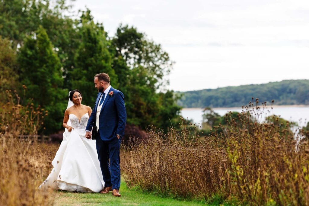 Beautiful outdoor wedding scene with a bride and groom holding hands in a natural setting by the lake, surrounded by greenery and wildflowers, capturing a special moment.