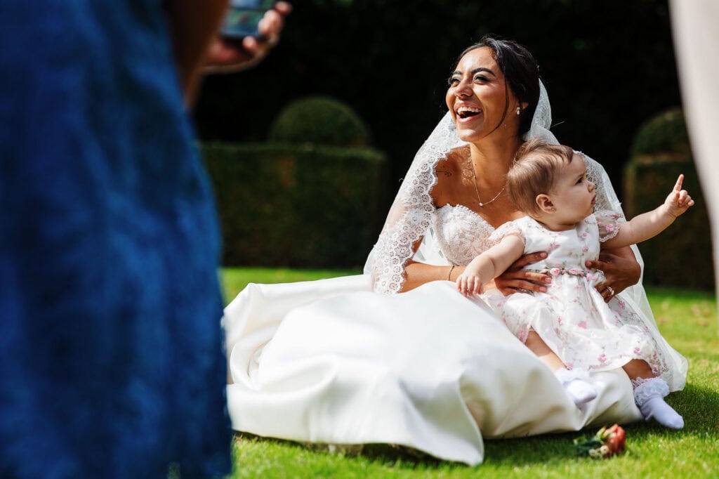 Happy bride in wedding dress holding a young girl outdoors, smiling and enjoying the moment at BGS Weddings, with lush green background.