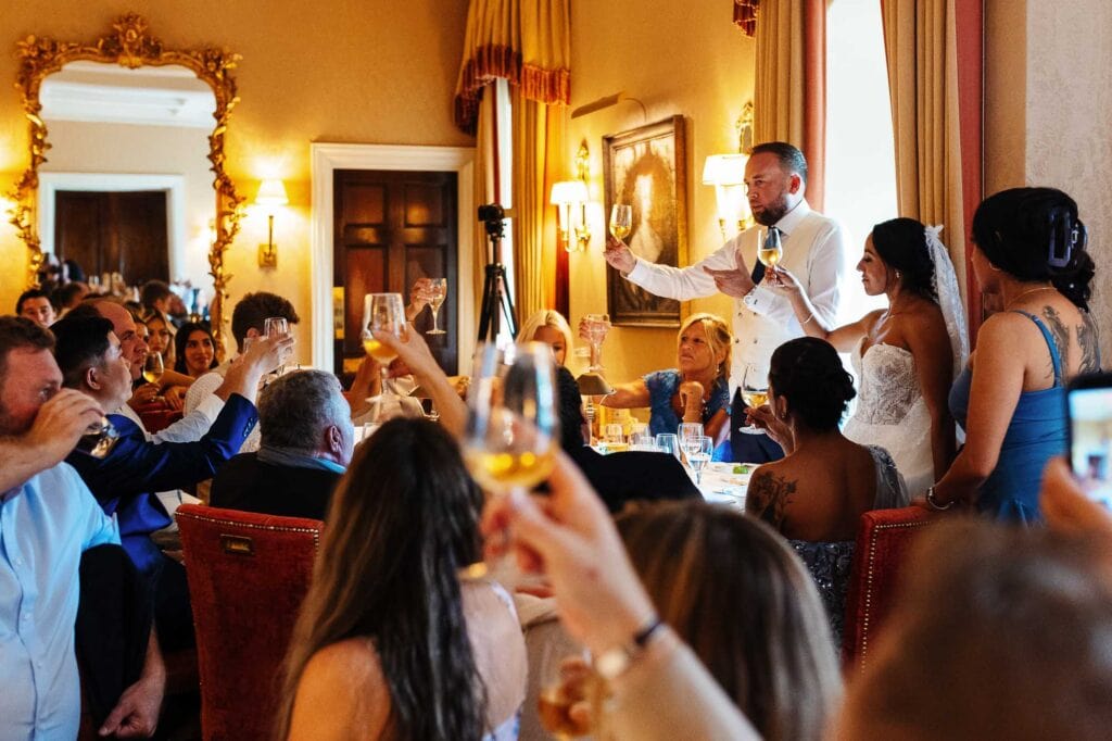 Wedding speech at BGS Weddings with bride, groom, and guests raising glasses in a luxurious room with ornate decor and warm lighting.