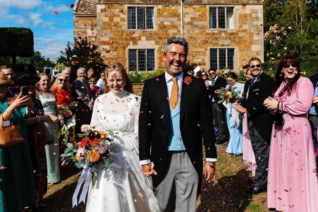 Happy bride and groom at BGS Weddings, surrounded by friends and family outdoors, with a historic stone building in the background, capturing a joyful moment.
